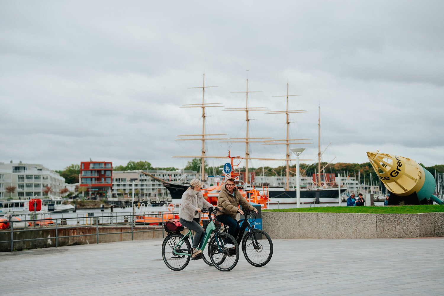 Zwei Personen auf Fahrrädern, eine Frau und ein Mann, stehen an einem Hafen mit Schiffen und einem großen gelben Objekt im Hintergrund. Es sind mehrere Boote und eine Hafenanlage sichtbar.